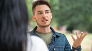 This image shows a young man, gesturing while having a conversation outdoors. If asked "How to answer why you're still single?", it's best to respond confidently, focusing on your values and personal journey without feeling pressured to justify your status.