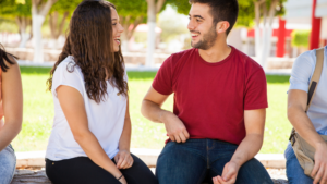 The photo shows a man and woman laughing together outdoors, creating a lighthearted moment. It raises the question, "How do you know if a girl is friendly or flirting?" The purpose of the image is to capture that subtle dynamic between casual friendliness and potential flirtation.