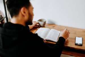 The image shows a man sitting at a wooden table, reading a book with his phone nearby. This visual represents the theme "How To Date As A Shy Guy?" by highlighting a moment of introspection and self-improvement, key steps for shy individuals to build confidence and overcome shyness in dating.