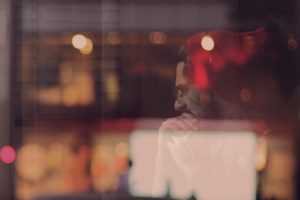 The image shows a man thoughtfully looking out a window, with city lights blurred in the background. This visual represents the theme "How To Date As A Shy Guy?" by illustrating a moment of deep contemplation and introspection, key aspects for shy individuals as they reflect on their feelings and work towards building the confidence needed for dating.