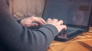 The image shows someone typing on a laptop, likely doing online research or interacting with others. It could represent investigating or testing online interactions, especially in the context of the question *"How Do You Test a Romance Scammer?"*, as the person might be trying to spot deceptive behavior or confirm suspicions.