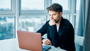 A man is focused on his laptop, possibly analyzing or investigating something important. This connects to how to outsmart a romance scammer, as he may be scrutinizing interactions or looking for signs of deception in an online relationship. The image emphasizes the careful process of protecting oneself from scams.