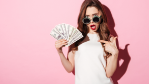 The image shows a young woman holding a fan of hundred-dollar bills, looking surprised and excited. She is wearing sunglasses and a white sleeveless top against a pink background, pointing at the money. The image emphasizes themes of wealth and financial success.