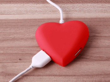 A red heart-shaped device connected by a white USB cable, symbolizing emotional connection and the idea of “recharging” love.