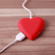 A red heart-shaped device connected by a white USB cable, symbolizing emotional connection and the idea of “recharging” love.