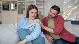 A man gently comforts a woman sitting beside him on a couch, showing concern while she looks distant. The indoor setting and soft body language highlight emotional connection, support, and empathy in relationships during vulnerable moments.