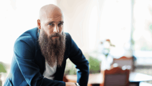 Bald man with a long beard wearing a navy blazer, leaning forward in a softly lit room—capturing quiet strength in dating as a quiet guy.