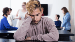A quiet, introverted young man sits alone at a desk while others chat in the background. The image illustrates the challenge introverts face in social environments and supports the topic how-introverts-talk-to-women by showing how isolation or overthinking can affect confidence.
