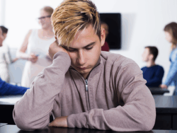 A quiet, introverted young man sits alone at a desk while others chat in the background. The image illustrates the challenge introverts face in social environments and supports the topic how-introverts-talk-to-women by showing how isolation or overthinking can affect confidence.
