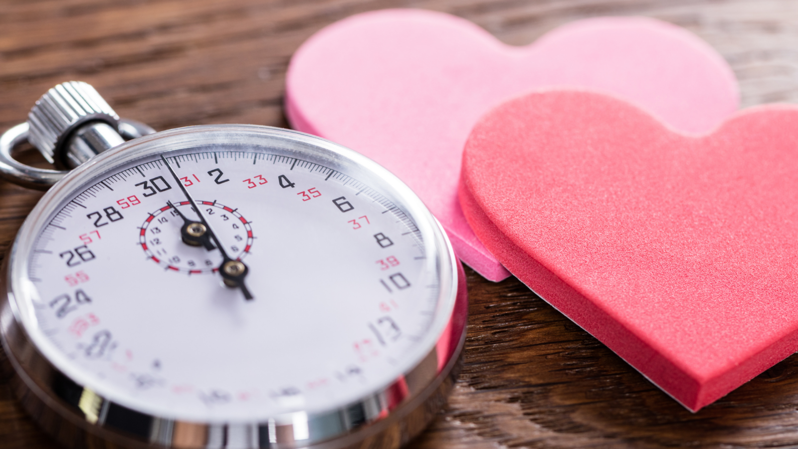 Silver stopwatch beside two paper hearts on a wooden table, illustrating the timed five-minute rounds of speed dating.